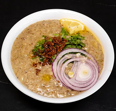 Bowl of haleem topped with fried onions, coriander, red onion rings and a lemon wedge