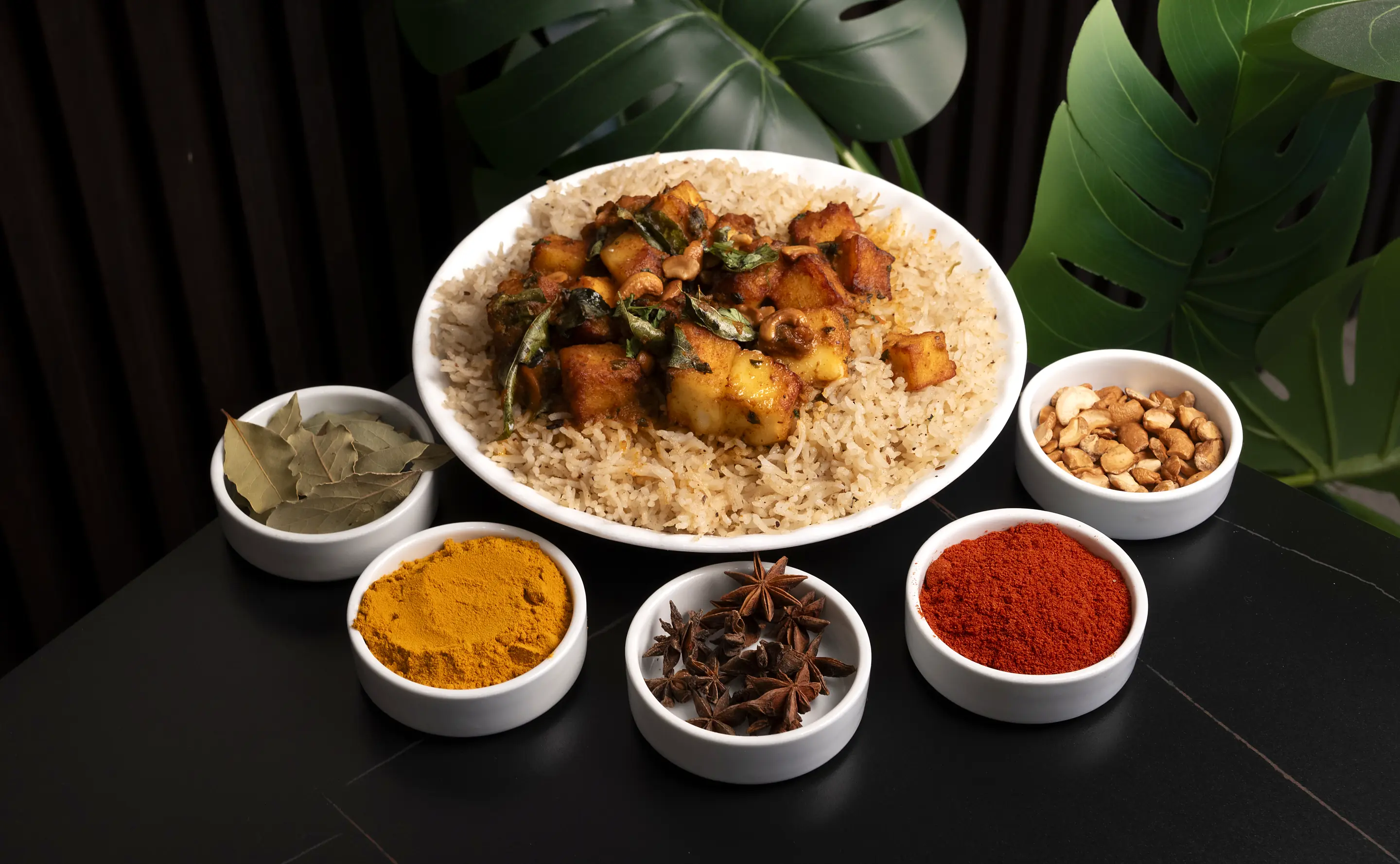 Plate of mandi rice topped with spiced paneer and curry leaves, with small bowls of bay leaves, turmeric, star anise, chilli powder and cashews