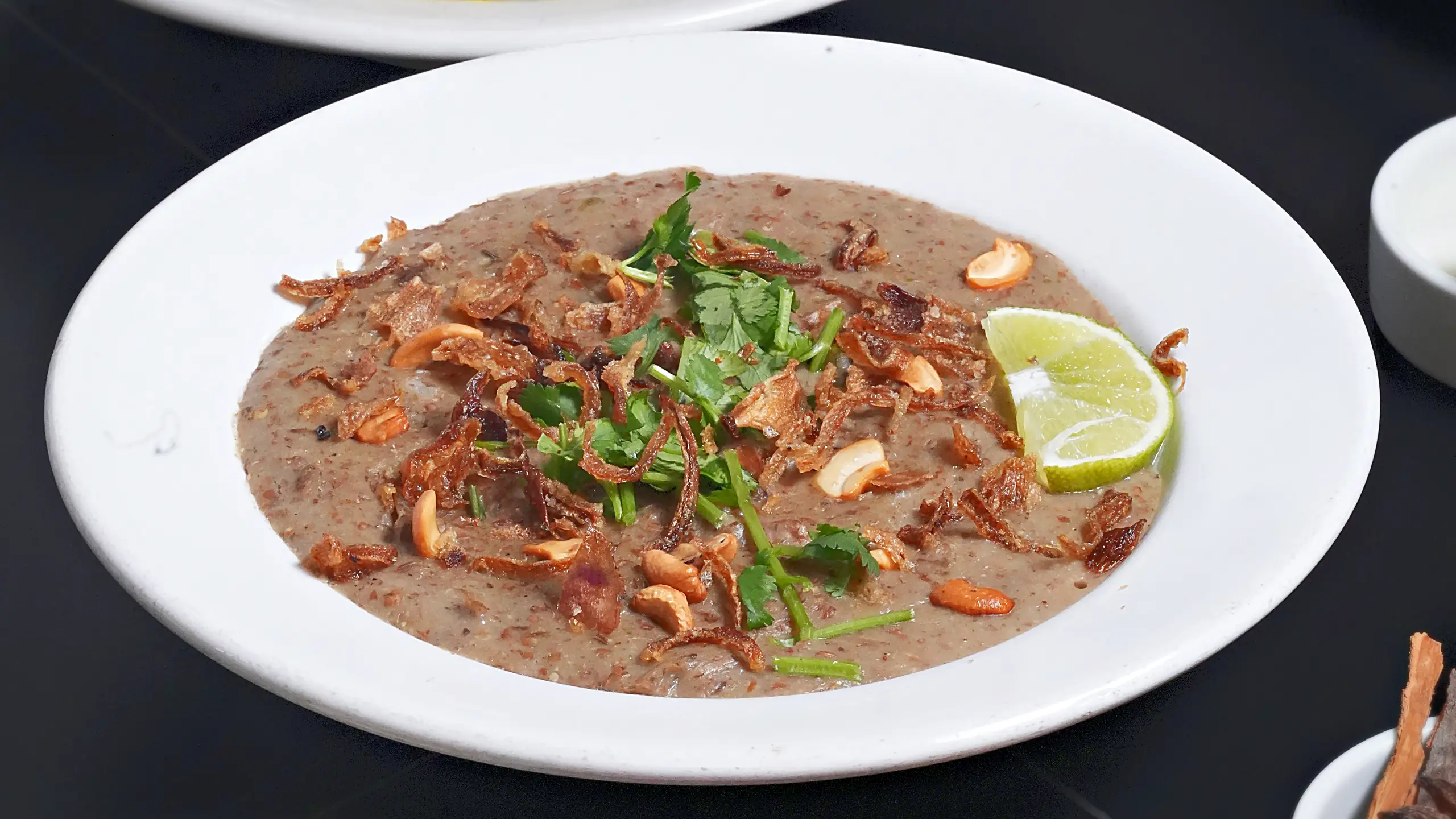 Bowl of haleem topped with fried onions, cashews, coriander and lime, beside a bowl of meat marag and small bowls of ground spices