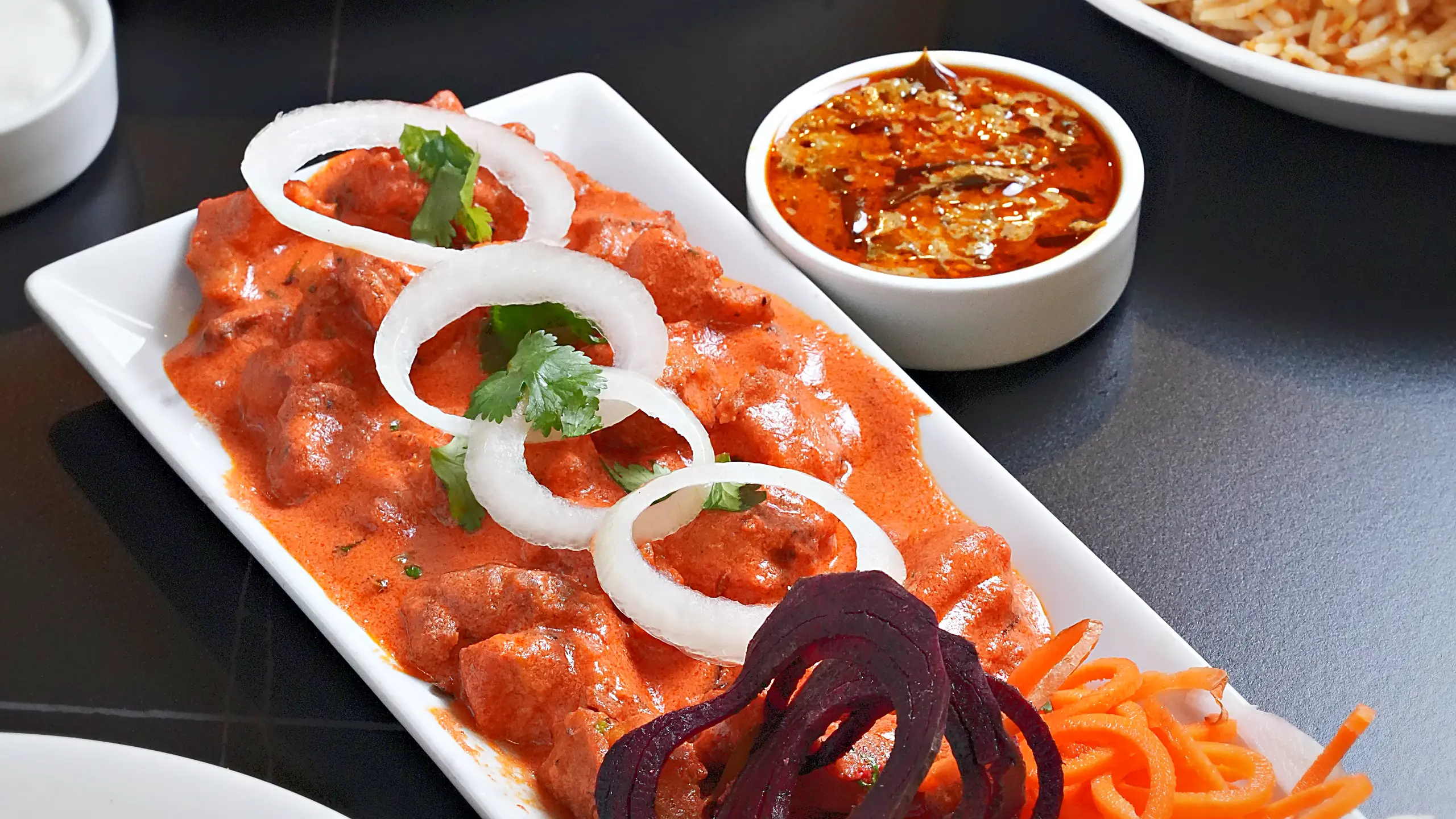 Chicken in creamy orange gravy with onion rings, beetroot and carrot garnish, next to biryani and a mandi platter